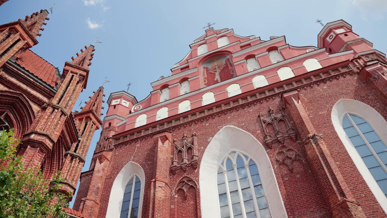 A ornamentada fachada gótica da Igreja Bernardina em Vilnius ergue-se majestosamente sobre exuberantes árvores verdes sob um céu azul claro, exibindo seus intrincados detalhes arquitetônicos sob a luz vibrante do sol