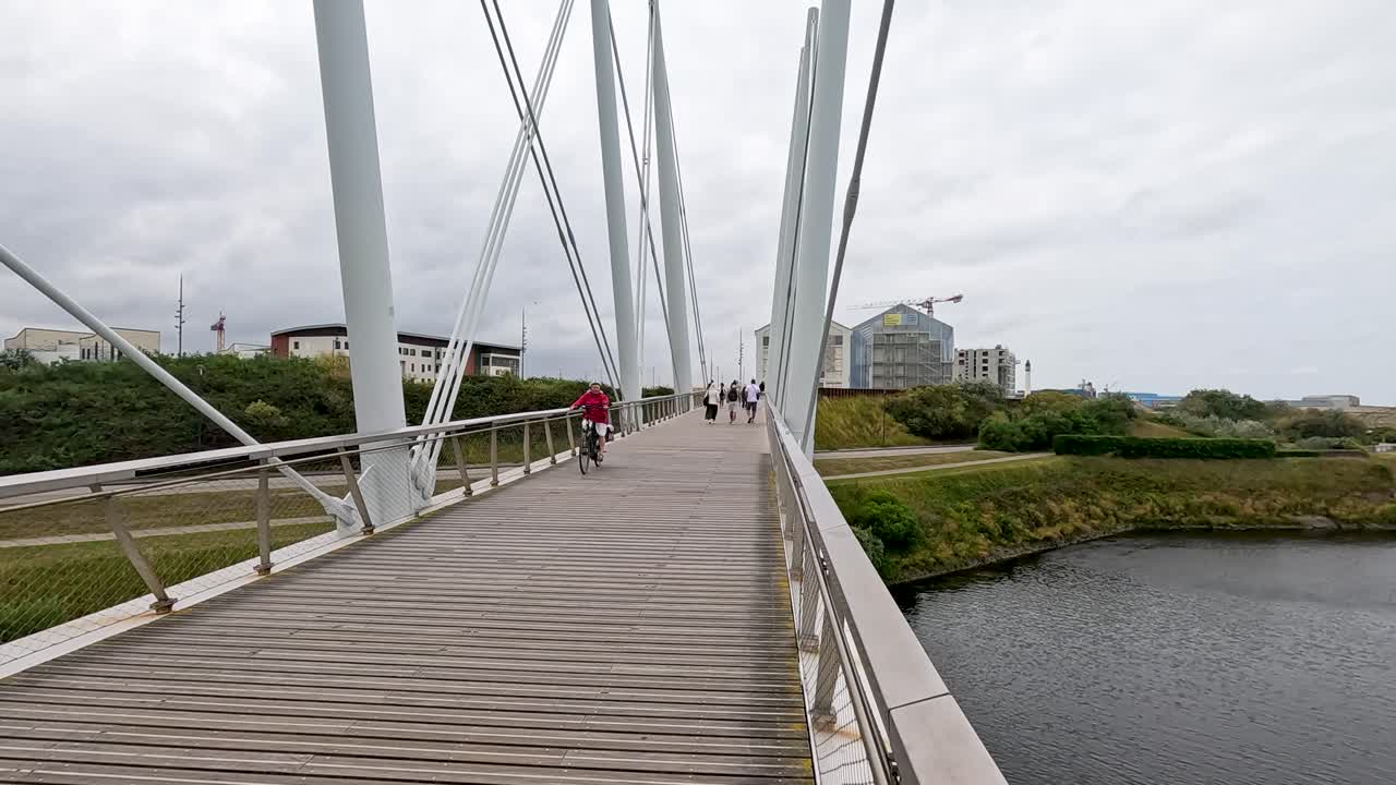 Two cyclists cross a modern pedestrian bridge in Dunkirk, France, on an overcast day