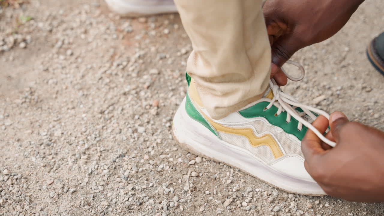 Primo piano delle mani di un padre nero che allaccia la scarpa da ginnastica del figlio sulla ghiaia. Pantaloni beige e scarpa verde-gialla. Cura delicata e movimento costante. Una routine mattutina prima di una passeggiata o di un gioco, un momento intimo di genitorialità che mostra.