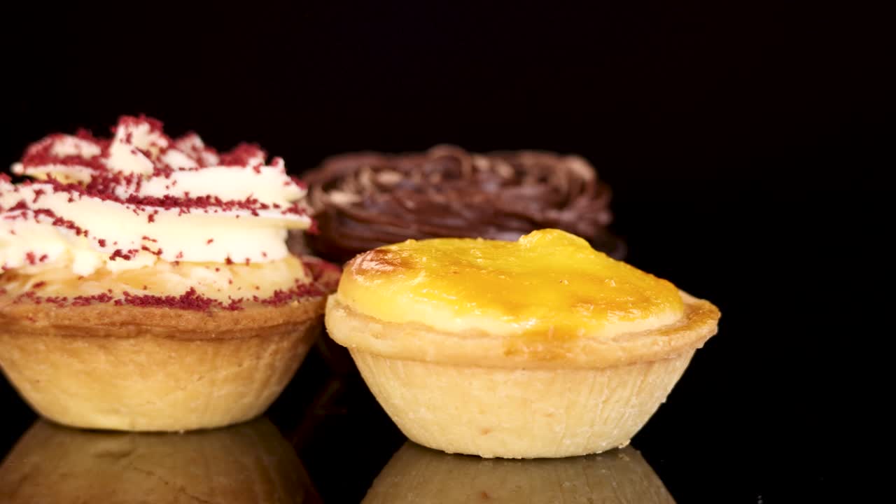 Three mini tarts—chocolate, red velvet, custard—rotate on glossy black surface under studio lighting