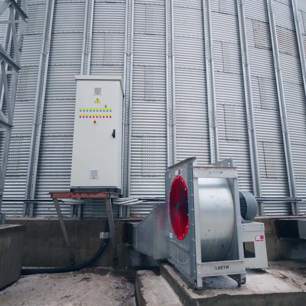 Big ventilators placed outside a huge cylinder of grain elevator. Movement from metal support near grain elevator to the back of one ventilator