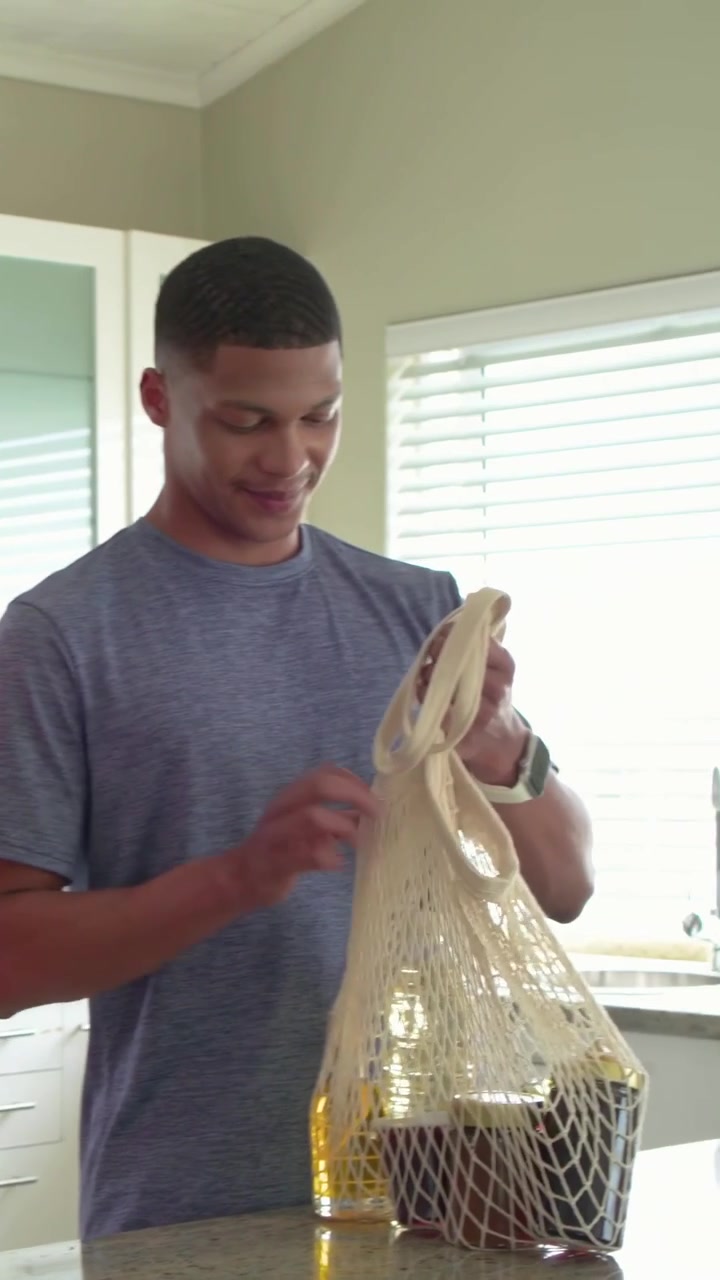 Vertical video: Entering woman with mesh bag nudging man unpacking groceries at kitchen, copy space