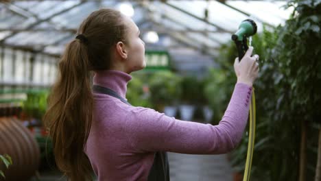 Young-smiling-female-gardener-in-uniform-watering-plants-with-garden-hose-in-greenhouse.-Slowmotion-shot