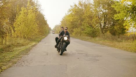 tracking shot of couple riding motorcycle on forest road in autumn. attractive young man in sunglasses driving his chopper while