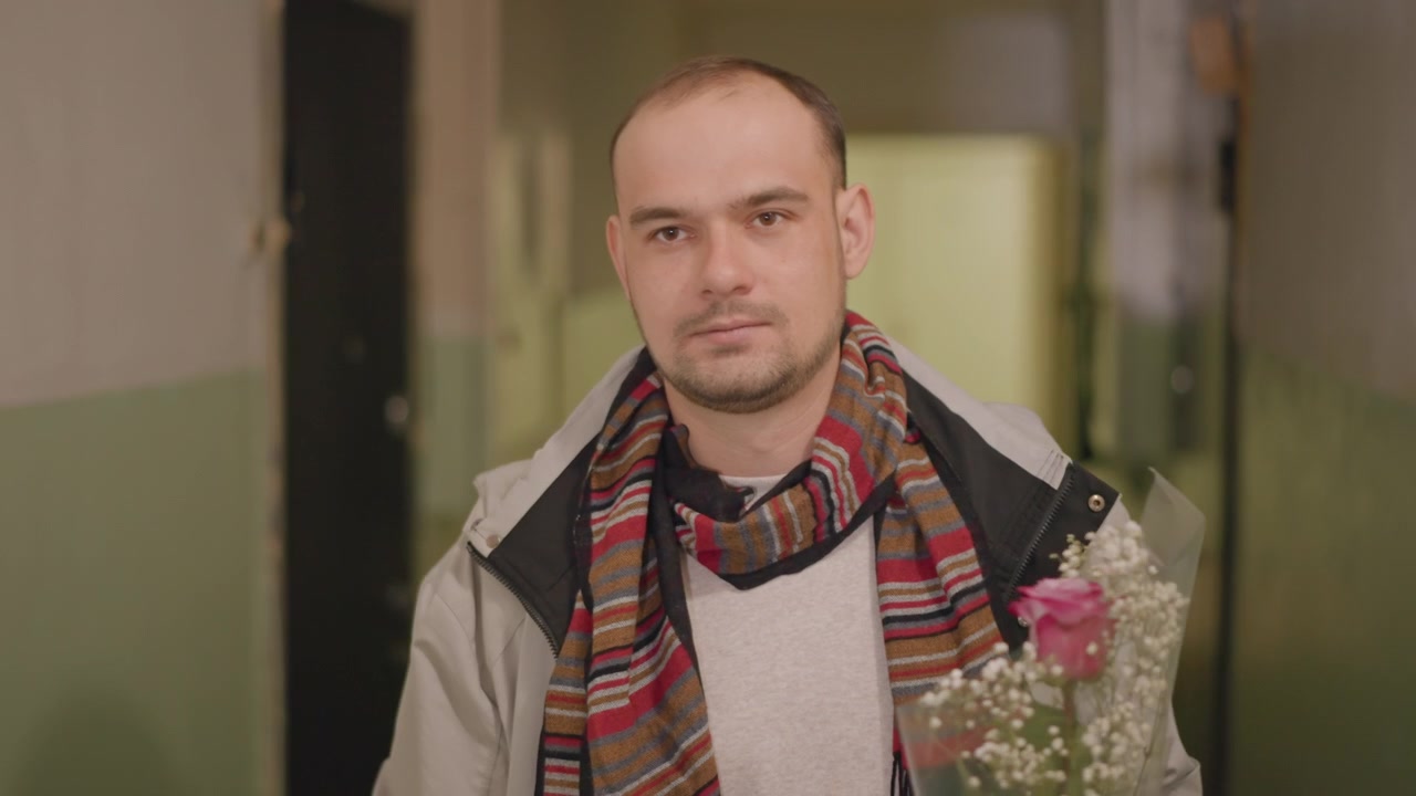 Un hombre de pie en un estrecho pasillo residencial, sosteniendo un ramo envuelto a la altura del pecho, mirando a la cámara con expresión tranquila, una sutil inclinación hacia adelante, luz cálida, puertas cerradas y un ambiente interior sereno.