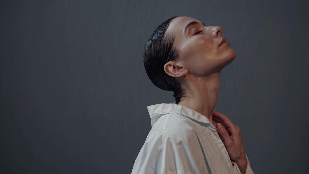 Young woman with wet hair tied in a braid meditates with closed eyes, wearing a white shirt, against a dark gray background, conveying a sense of tranquility and mindfulness