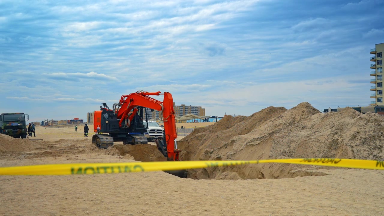 New York, USA, 1 April 2026: Orange excavator digging deep trench on sandy beach shore. Heavy machinery excavating sand on a beach with urban buildings and yellow caution tape in the foreground.