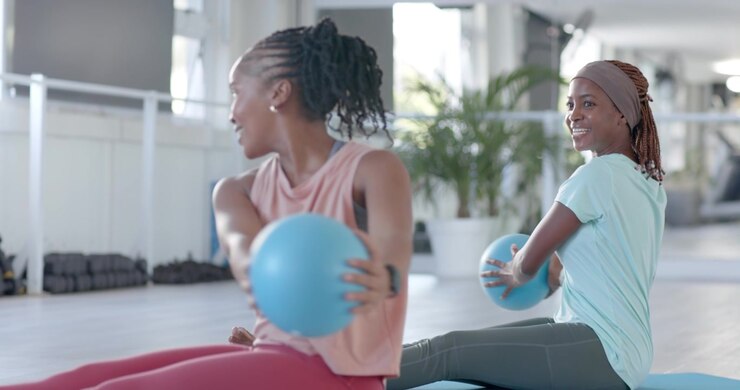 African American female friends passing med ball after front woman lifting it, training core in gym