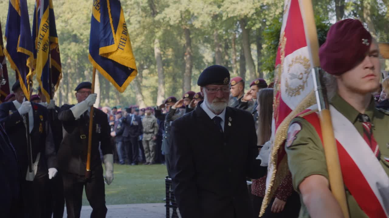 Veterans in maroon berets march with ceremonial flags at Oosterbeek War Cemetery, paying respects during a solemn memorial event for fallen soldiers of WWII. Oosterbeek, Gelderland