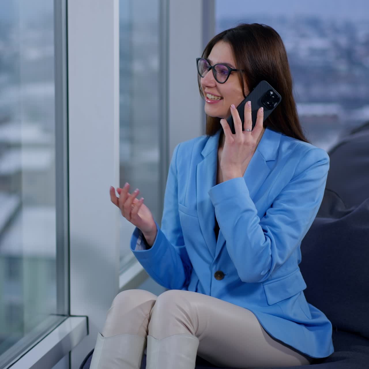 Successful resilient business lady sits in office speaking on the phone. Brunette woman in blue jacket and glasses looks at window having conversation and gesturing with her hand