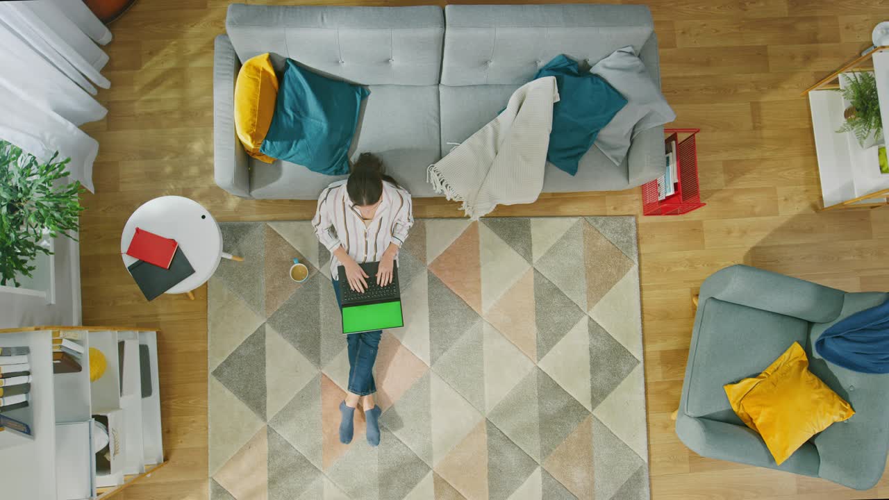 Una giovane ragazza è seduta per terra, beve caffè e usa un laptop con schermo verde. Accogliente soggiorno con interni moderni, con tappeto, divano, sedia, tavolo, scaffale, pianta e pavimento in legno. Vista dall'alto