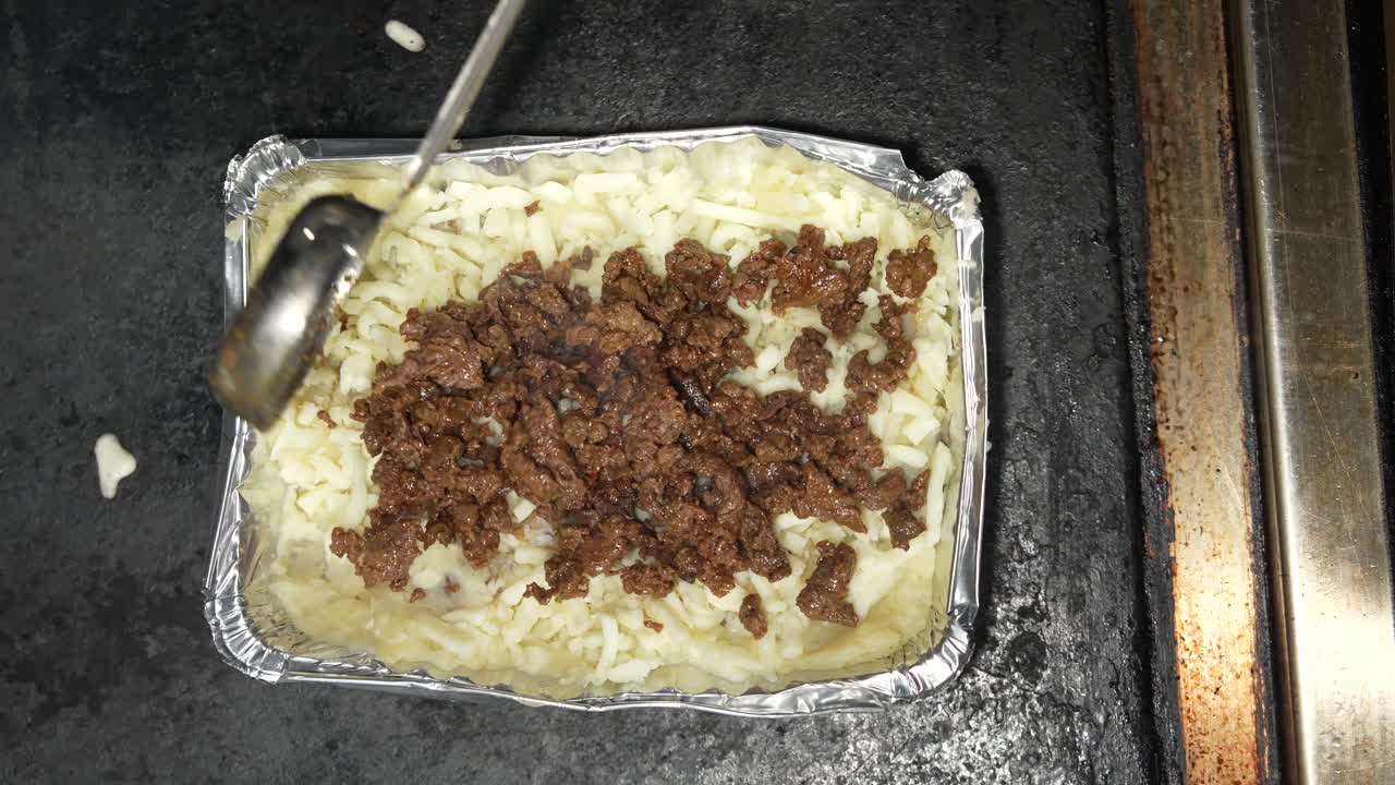 Video en primer plano de carne asada recién hecha siendo servida sobre papa loca con patatas y queso fundido en una bandeja de aluminio. La acción enfatiza la preparación fresca, la comida para llevar y la cocina callejera.