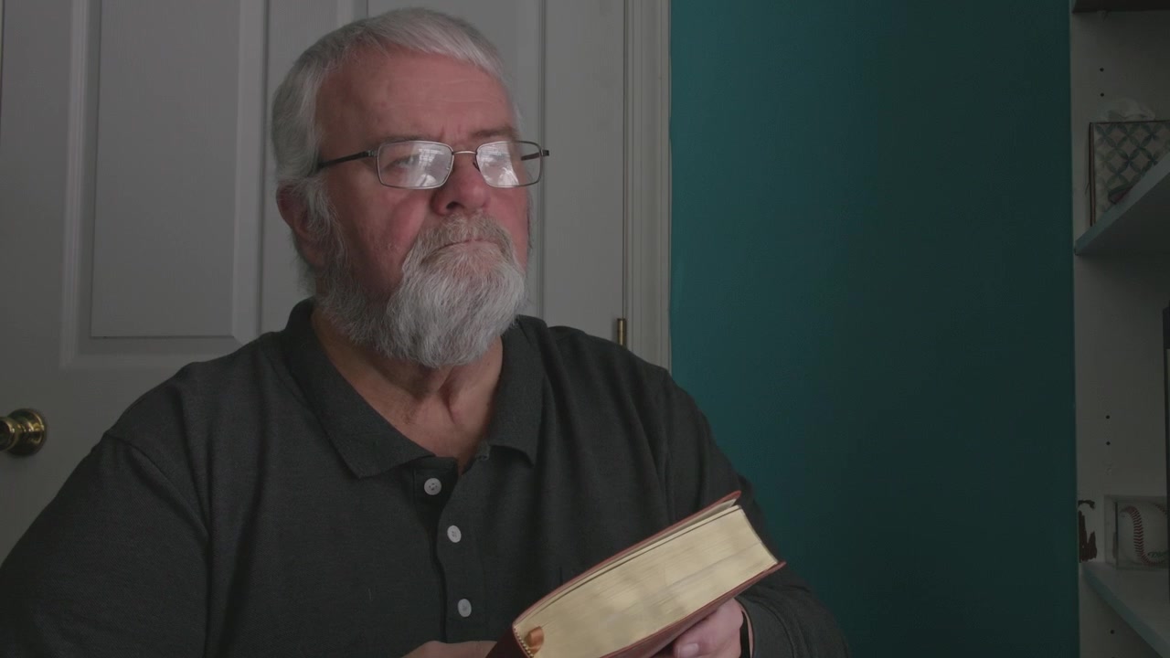 A man with a beard reads a book in a bright room. He sits quietly and focuses on the pages. Sunlight enters through the window. This takes place in the afternoon.