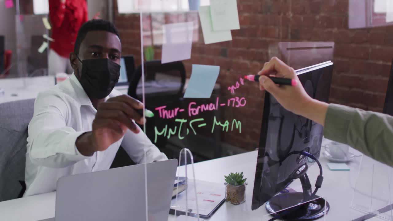 Divers male business colleagues wearing masks sitting at desks using glass wall