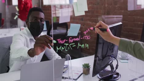 Divers-male-business-colleagues-wearing-masks-sitting-at-desks-using-glass-wall