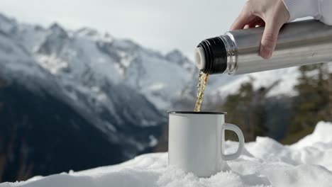 Verter-Café-Líquido-Caliente-De-Un-Termo-En-Una-Taza-Durante-Un-Invierno-Frío-Con-Montañas-Llenas-De-Nieve-Como-Fondo