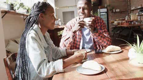 feliz pareja afroamericana hablando y bebiendo café en la mesa en una cafetería soleada, cámara lenta