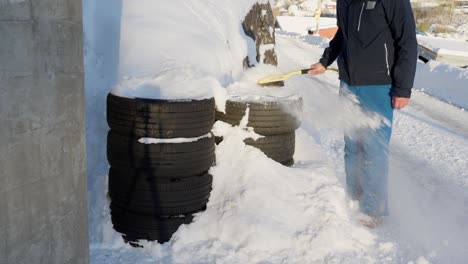 man brushes snow off summer tyres left outside in two piles near a garage, showing deterioration during snowy winter conditions