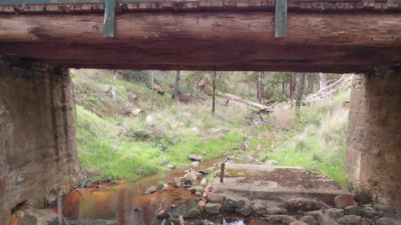 drone vuela hacia y debajo de un puente de madera y piedra sobre un arroyo, con un cobertizo de madera a la izquierda, en la selva australiana