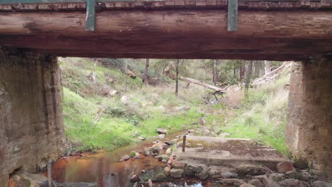 Drone-Vuela-Hacia-Y-Debajo-De-Un-Puente-De-Madera-Y-Piedra-Sobre-Un-Arroyo,-Con-Un-Cobertizo-De-Madera-A-La-Izquierda,-En-La-Selva-Australiana