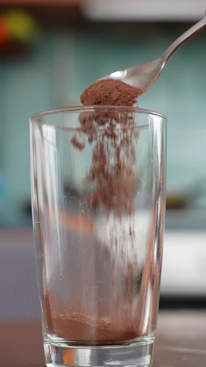 Primo piano verticale di cacao in polvere che cade da un cucchiaio in un bicchiere alto vuoto su un tavolo di legno, per iniziare una ricetta di bevanda al cioccolato fatta in casa in una cucina luminosa