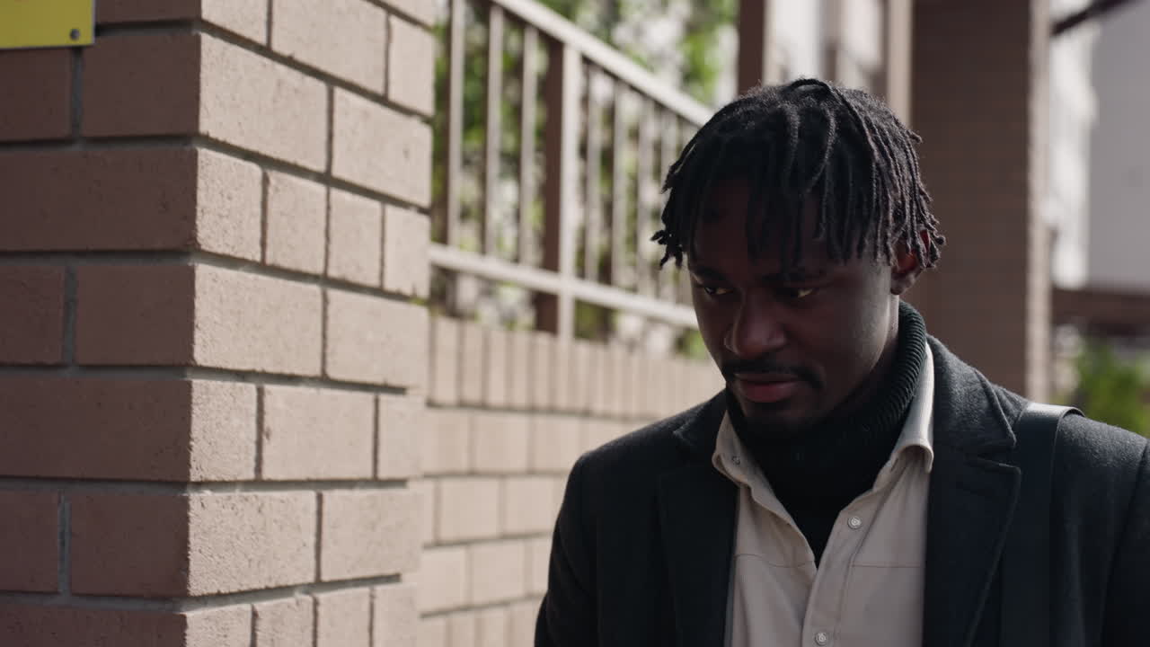 African man in coat walks beside brick railing while glancing down at phone, confirms meetup message and continues toward friend, soft afternoon sun, muted tones, casual stride, focused mood