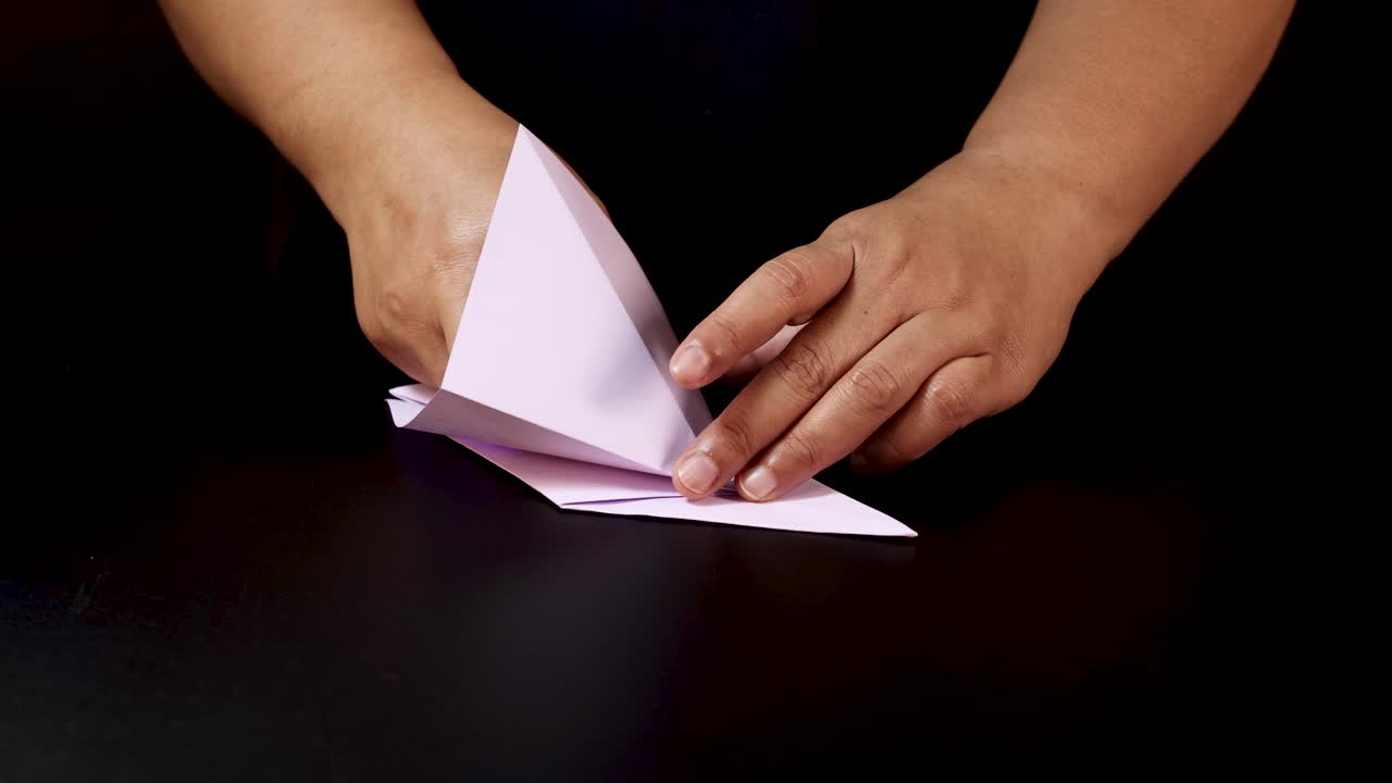 Person folds pink paper on black table, step-by-step, under soft studio lighting, overhead view