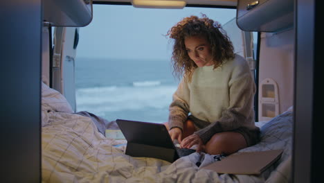 camp traveller holding cellphone sitting trailer bed with laptop. woman surfing