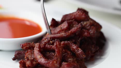 close-up of fried pork slices being dipped into a vibrant red sauce, showcasing thai cuisine in natural lighting