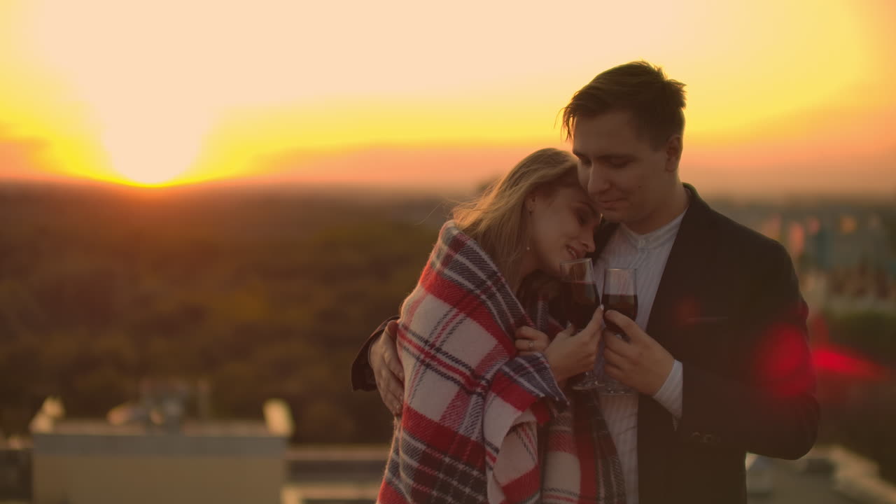Man and young pretty woman sitting on couch holding wine glasses and kissing on rooftop terrace at sunset