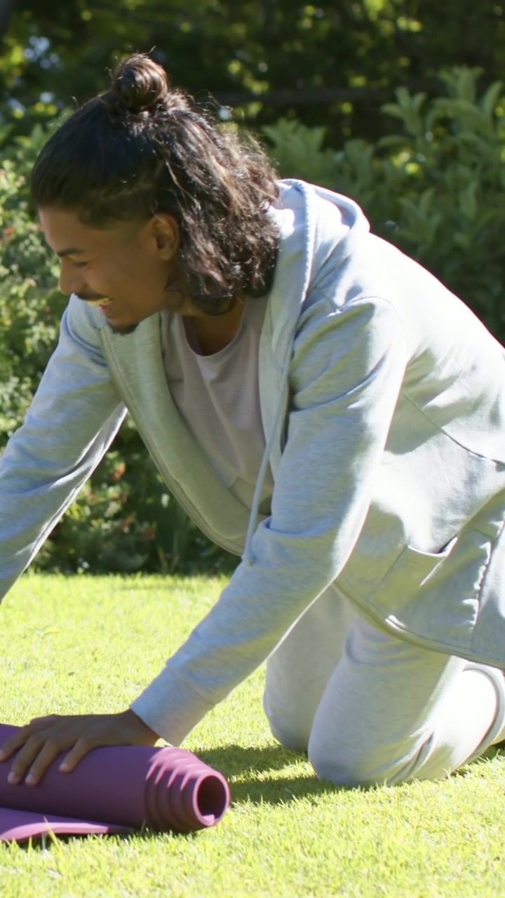 Video vertical: Hombre arrodillado tirando de una esterilla morada en un césped soleado, con una sudadera con cremallera, preparándose para hacer yoga