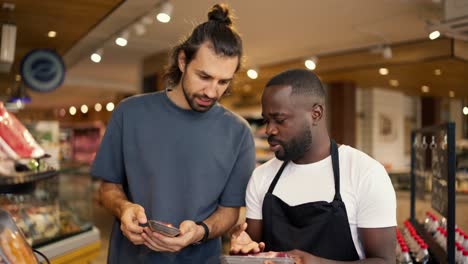 a-man-with-Black-skin-color-in-a-white-t-shirt-and-a-black-apron-helps-a-customer-of-a-supermarket-to-choose-a-product