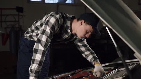 car service, repair, maintenance and people concept - mechanic man wearing work robe and cap with gloves on his hands inspecting all the mechanisms and their proper functioning.