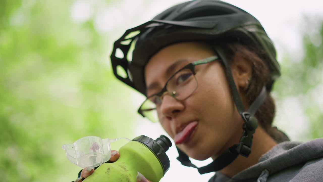 Ciclista asiatico, primo piano, beve da una bottiglia d'acqua, casco e occhiali visibili, lingua buffa ed espressione soddisfatta, sfondo verde sfocato, momento di idratazione fondamentale durante la corsa