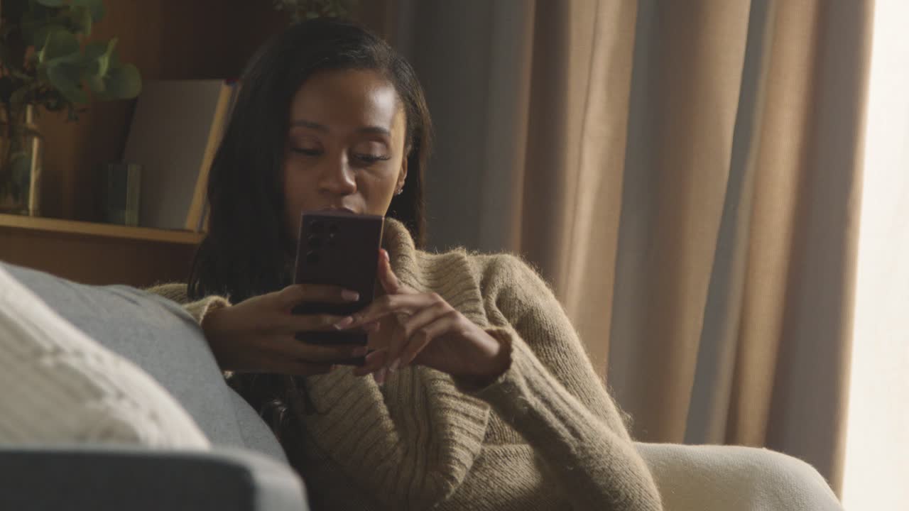 mujer joven relajándose en casa sentada en el sofá desplazándose por el teléfono móvil