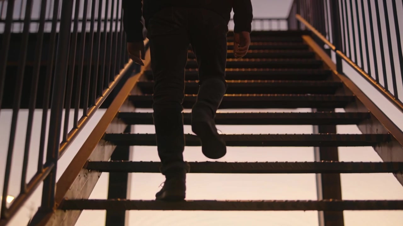 Lifting foot starting climb man walking up fire escape stairs to roof at sunset, rails, jacket