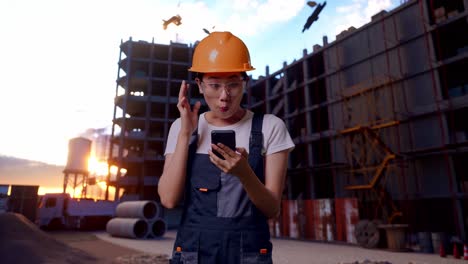 asian woman worker wearing goggles and safety helmet looking at smartphone, smiling, and saying wow while standing at construction site