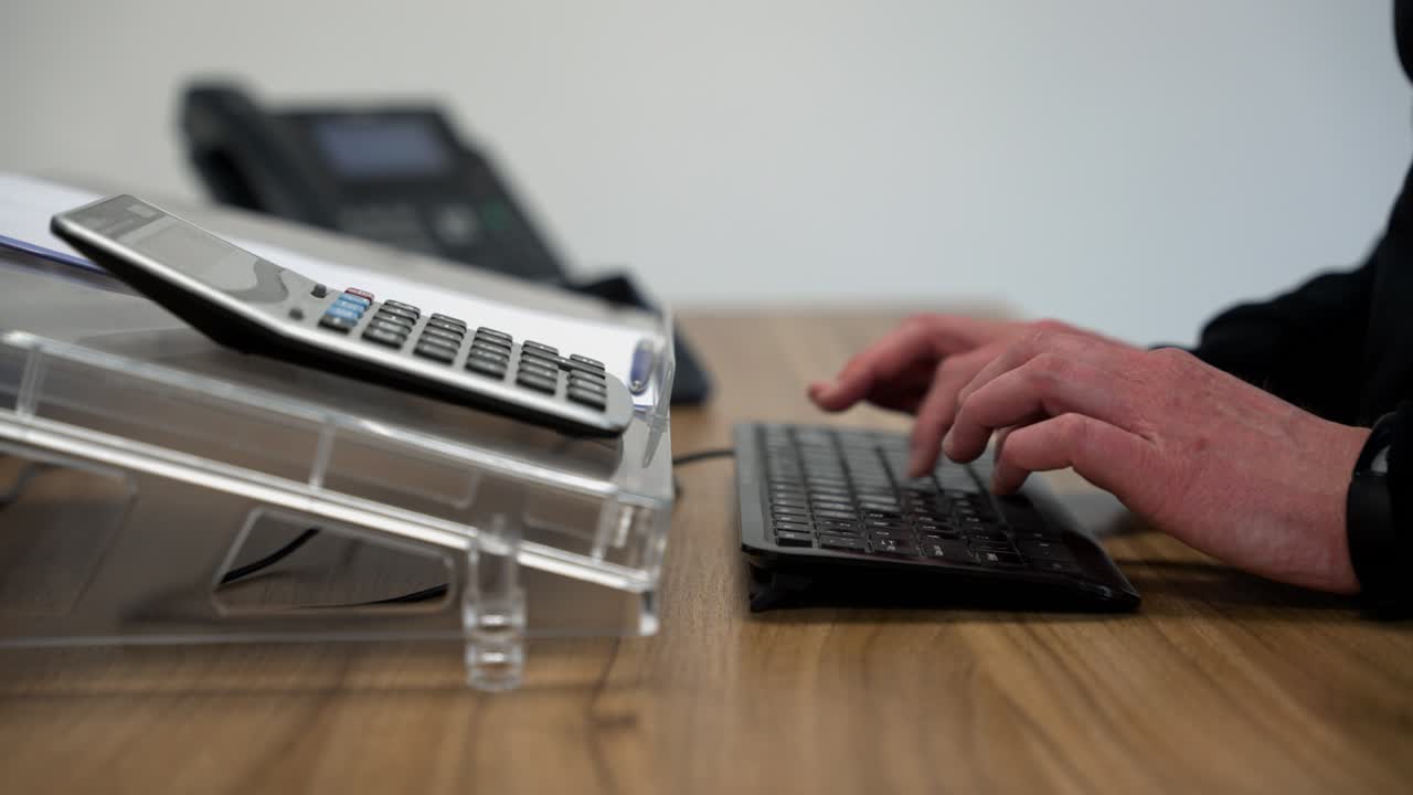 oficina trabajando en un escritorio de madera con paredes blancas escribiendo en el teclado en la oficina de ventas con una camiseta de manga larga y teléfono en el fondo, escribiendo en cámara lenta con un ligero movimiento lateral