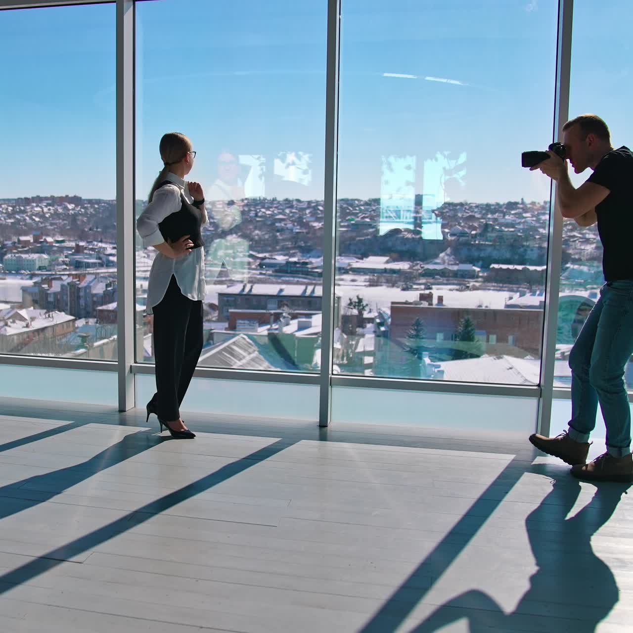 Attractive business woman poses to a photographer. Young female in glasses in black and white clothes posing on camera near large window indoors