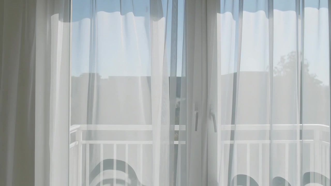 Tende velate che ondeggiano muovendosi attraverso le porte-finestre in vetro di una stanza, la brezza rivela la ringhiera del balcone.