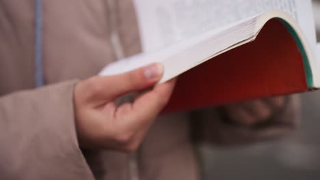 close-up view of lady's hands gently flipping pages of red book in autumn setting, blurred background with soft coat fabric visible, capturing peaceful reading moment in natural outdoor light