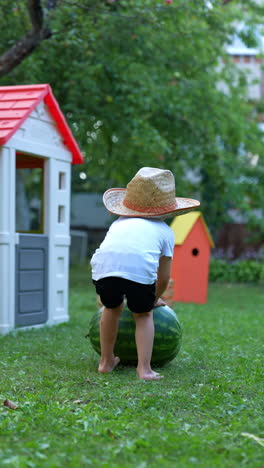 little baby wearing big straw hat comes up to a watermelon lying on the grass. unrecognized kid pushes the watermelon to the playhouse. vertical video.
