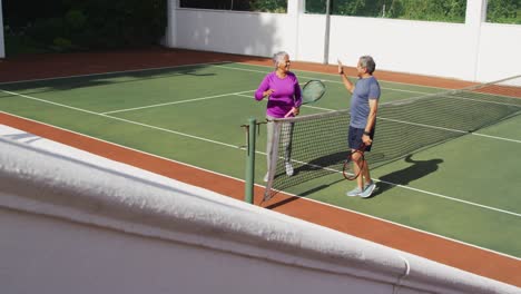 Video-of-happy-biracial-senior-couple-clapping-hands-after-match-on-tennis-court