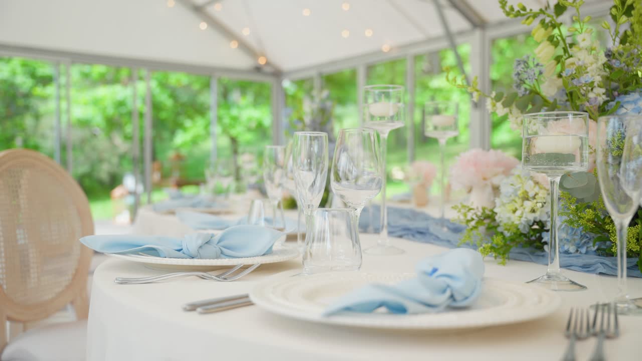 Elegant bryllupsreceptionsbord dekoreret med blå servietter, krystalglas, blomsterarrangementer og lysestager under et telt med naturligt grønt i baggrunden