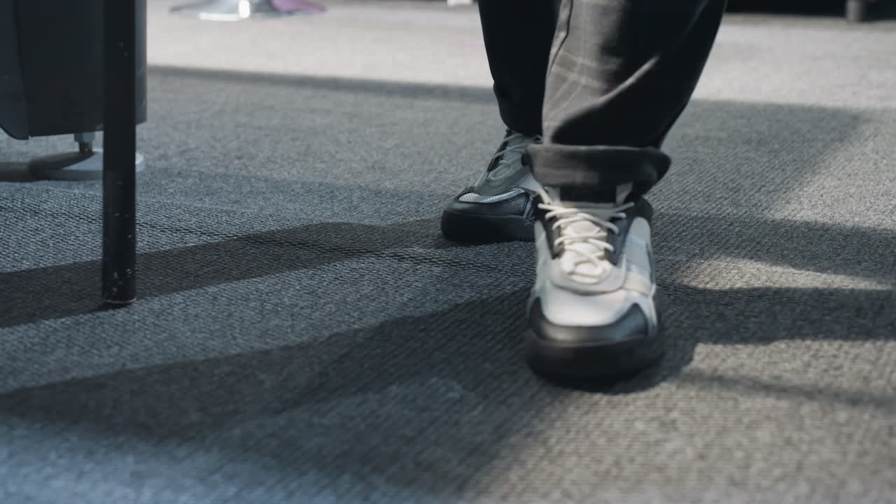 검은색 바지와 스니커를 입고 카으로 된 바닥을 고있는 남성 인물의 클로즈업 그림자와 움직임은 움직임을 강조합니다. 현대적인 신발과 캐주얼 스타일이 눈에 띄고 역동적인 조명이 있습니다.