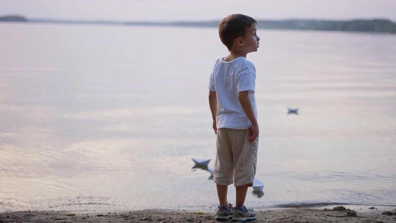 Tampak belakang anak laki-laki melihat perahu kertas di danau pada malam hari. Anak berdiri di tepi berpasir dengan perahu origami mengambang di air tenang yang memantulkan cahaya.