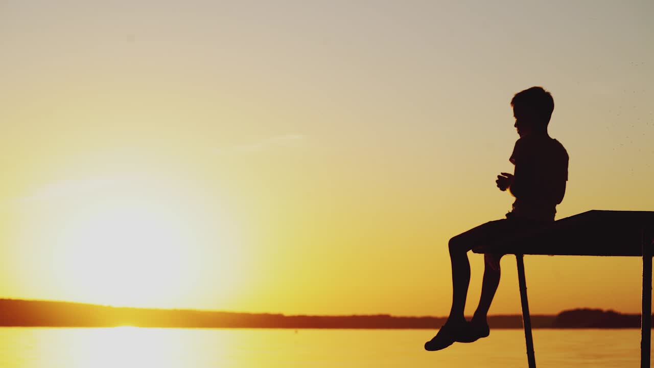 A little child is sitting on the bridge above the lake and throwing stones into the water at sunset in the summer on a warm evening.