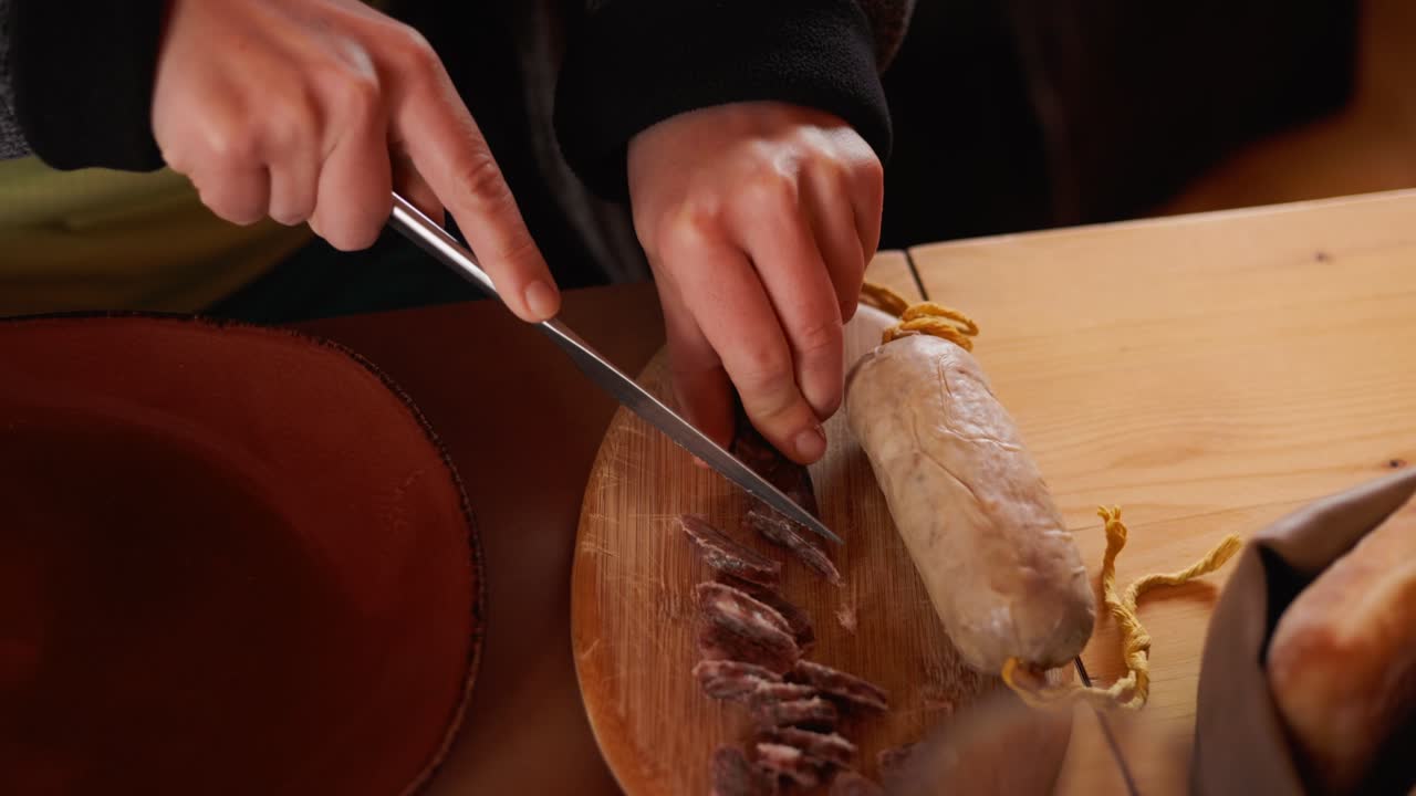 Hands slicing sausage with knife on wooden board. Plate visible beside. Detailed food preparation close-up.