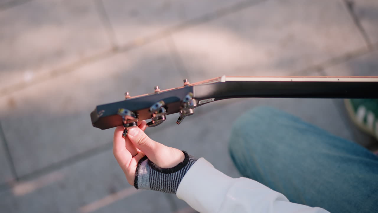 primo piano di artista di strada caucasico che modifica le chiavi di accordatura della chitarra sotto una luce solare soffusa, guanto senza dita e denim visibili, angolazione dall'alto della panchina, messa a punto rapida prima di suonare per strada, scena urbana spontanea, energia lofi allegra