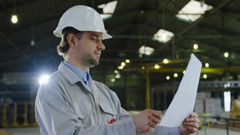 caucasian engineer wearing helmet and looking around and at blueprint in a big factory